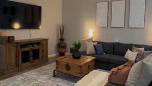 Een woonkamer met een donkere bank, een houten salontafel, een flatscreen televisie en een kamerkleed met een patroon.