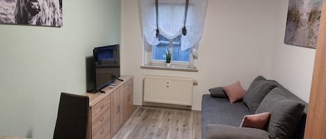 Ein kleiner, gemütlicher Raum mit einer Couch, einem Fernseher, einem Holzschrank und einem Fenster mit Vorhängen.