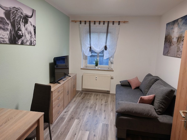 Ein kleiner, gemütlicher Raum mit einer Couch, einem Fernseher, einem Holzschrank und einem Fenster mit Vorhängen.