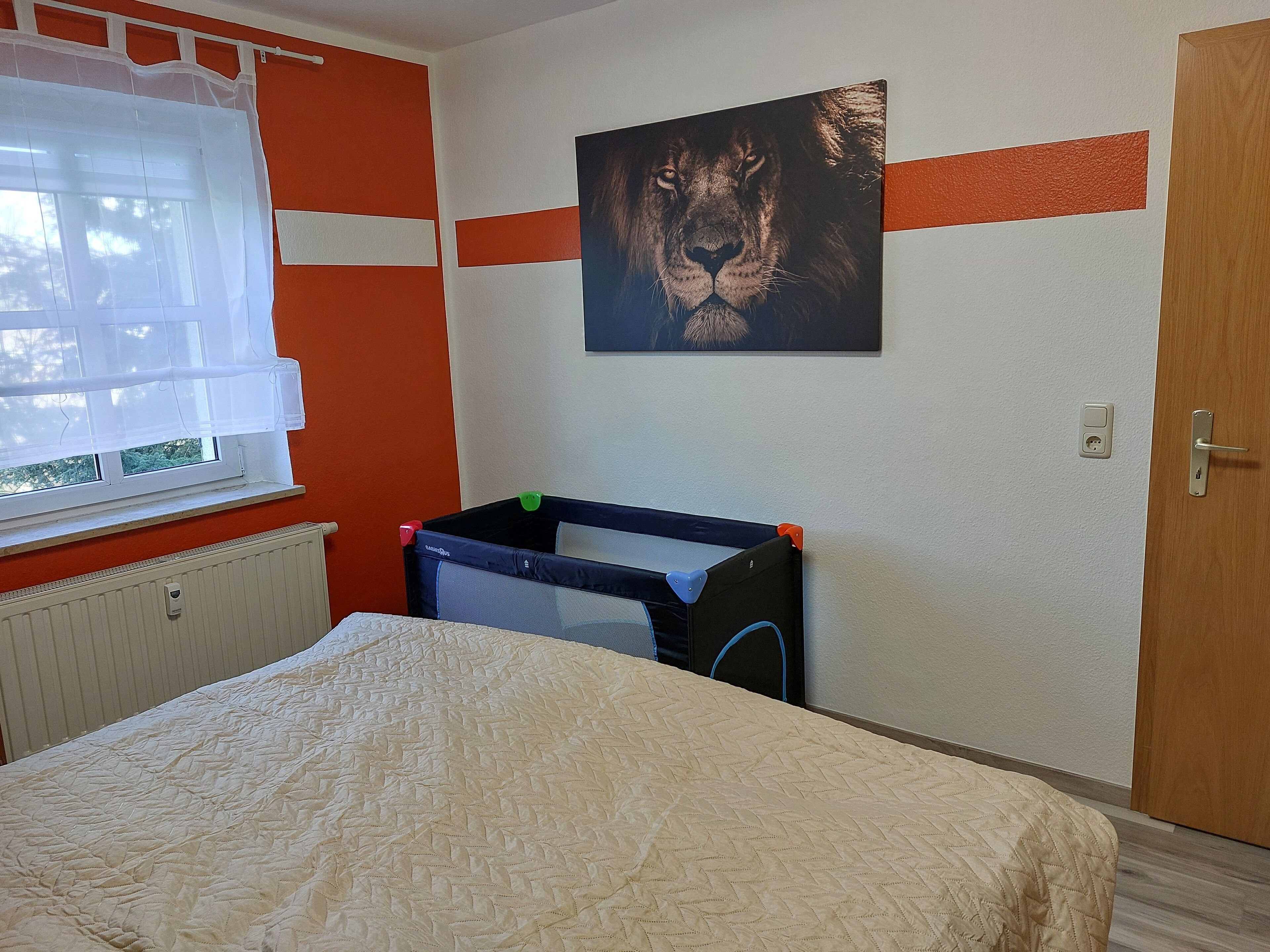 Ein Schlafzimmer mit einem Bett, einem Fenster mit weißen Vorhängen, einem Heizkörper, einem Wandbild eines Löwen und einer Tür.