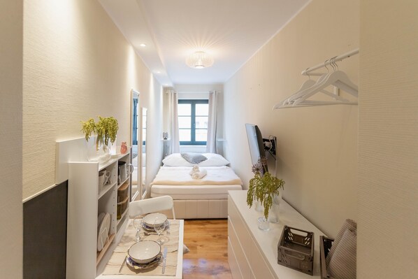 A narrow hotel room with a bed, a dining table set with plates, glasses, and cutlery, a small seating area, a TV, and a wall-mounted drying rack.