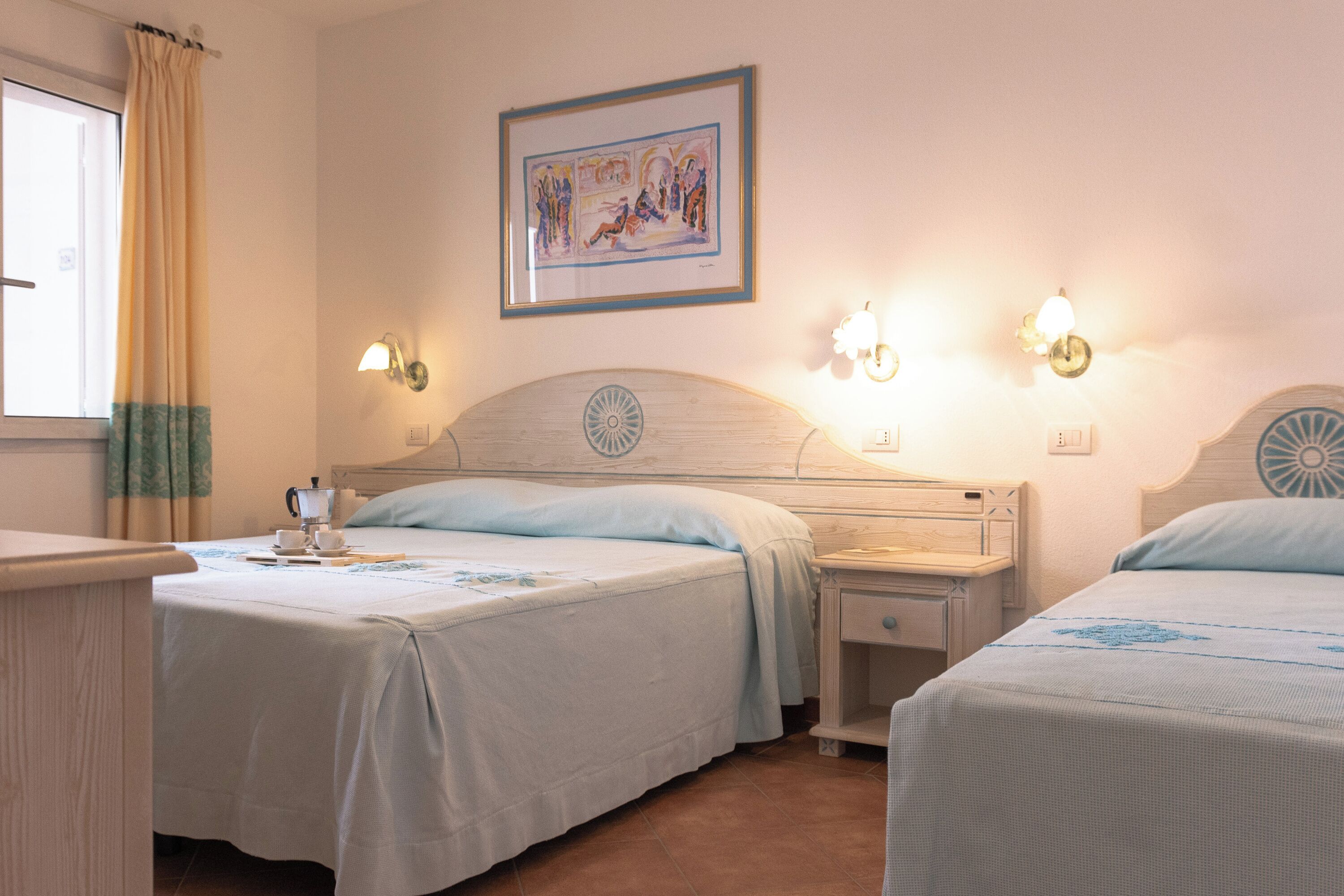 A hotel room with two beds, a nightstand, a bedside table, a window with curtains, and a wall-mounted painting.