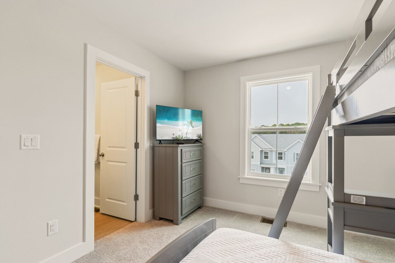 A bedroom with a bunk bed, a dresser, a television, and a window with a view of houses.
