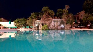 Ein Schwimmbecken mit Wasserfall, Liegestühlen und viel Grün.