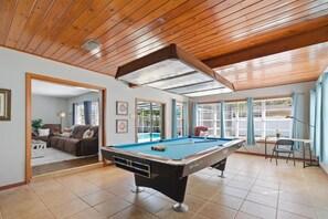 Ein Billardtisch in einem Raum mit Holzbalken, großen Fenstern und Blick auf einen Pool.