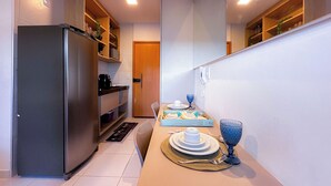 A narrow hallway with a refrigerator, shelves, a door, and a dining setup with plates, a bowl, and a glass. - Flat em resort - Várias Piscinas (Ipojuca)