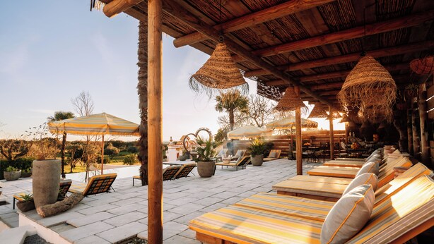 A sunlit outdoor lounge area with wooden furniture, striped cushions, and hanging woven lamps. - Herdade dos Moreiros (Arronches)