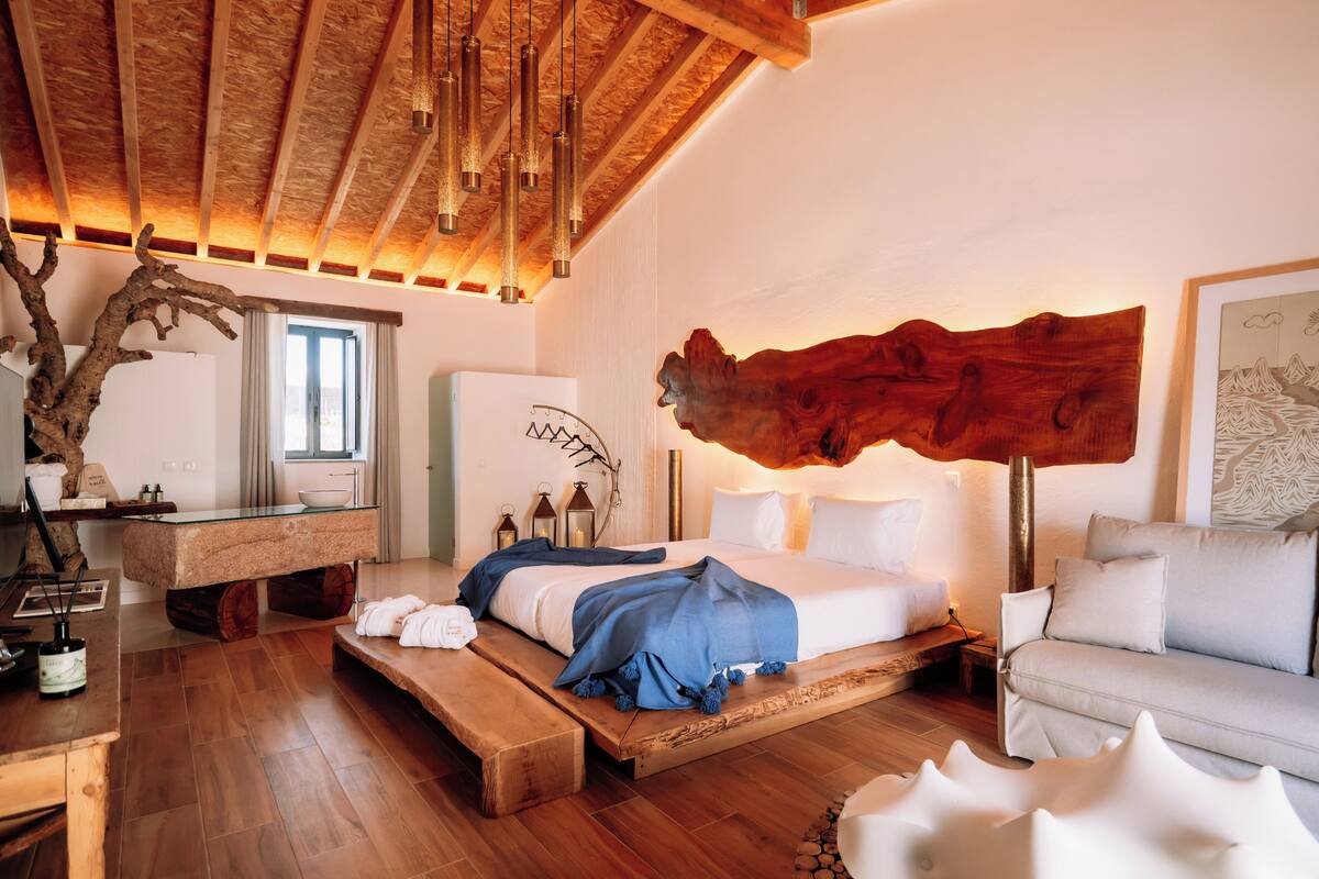 Un dormitorio con techo de madera, una cama con ropa de cama blanca, un banco de madera, un sofá blanco y una escultura de madera montada en la pared.