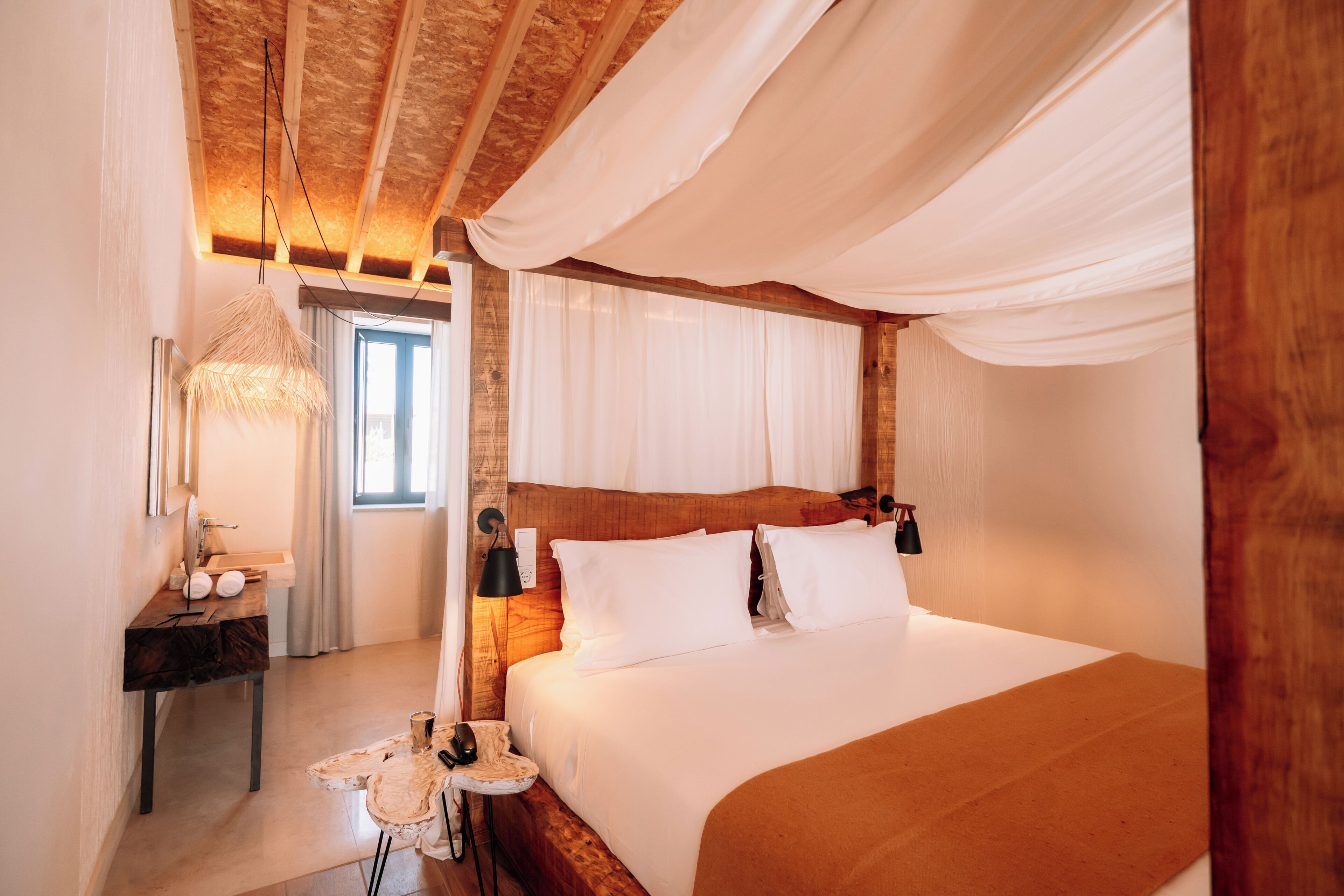 Un dormitorio con cabecera de madera, cama con ropa blanca, mesita de noche con lámpara y ventana con cortinas.