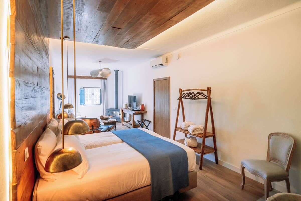 Un dormitorio con cabecera de madera, cama con manta azul, escritorio con silla y una silla mecedora de madera.