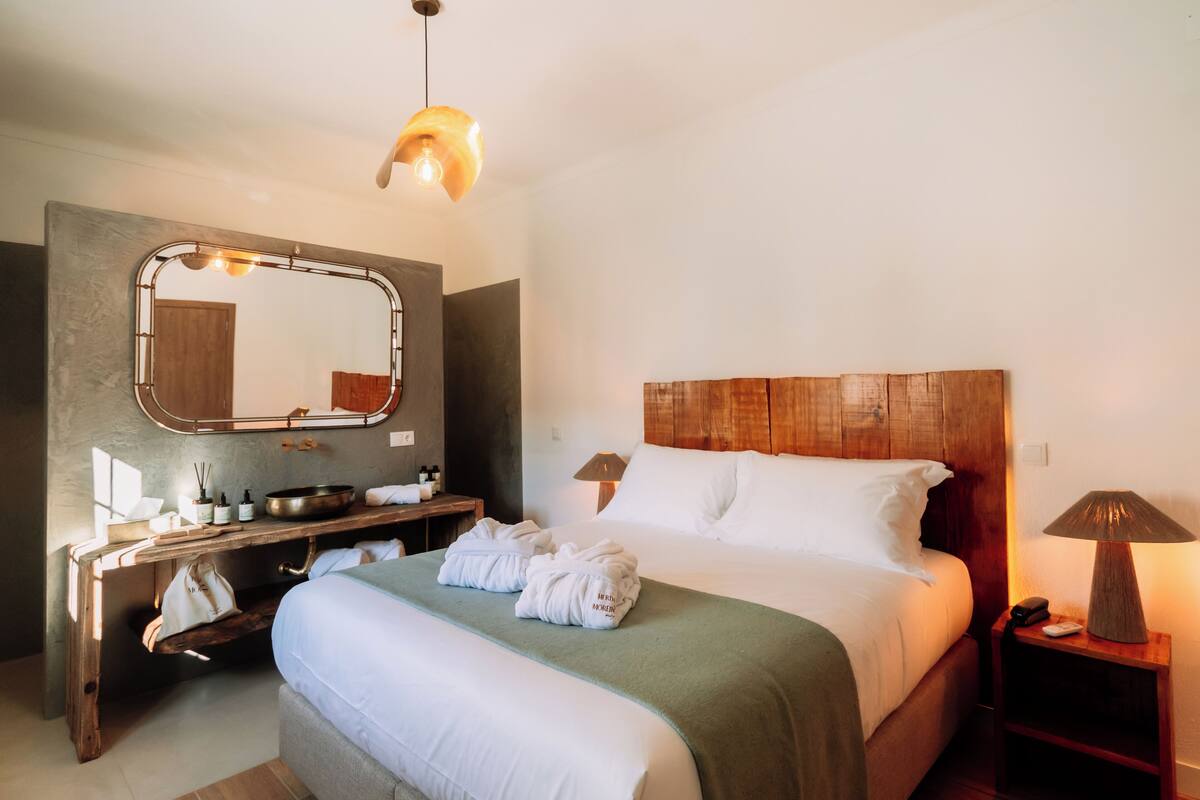 Habitación de hotel con cama, cabecera de madera, tocador con espejo, lámpara de noche y perchero.