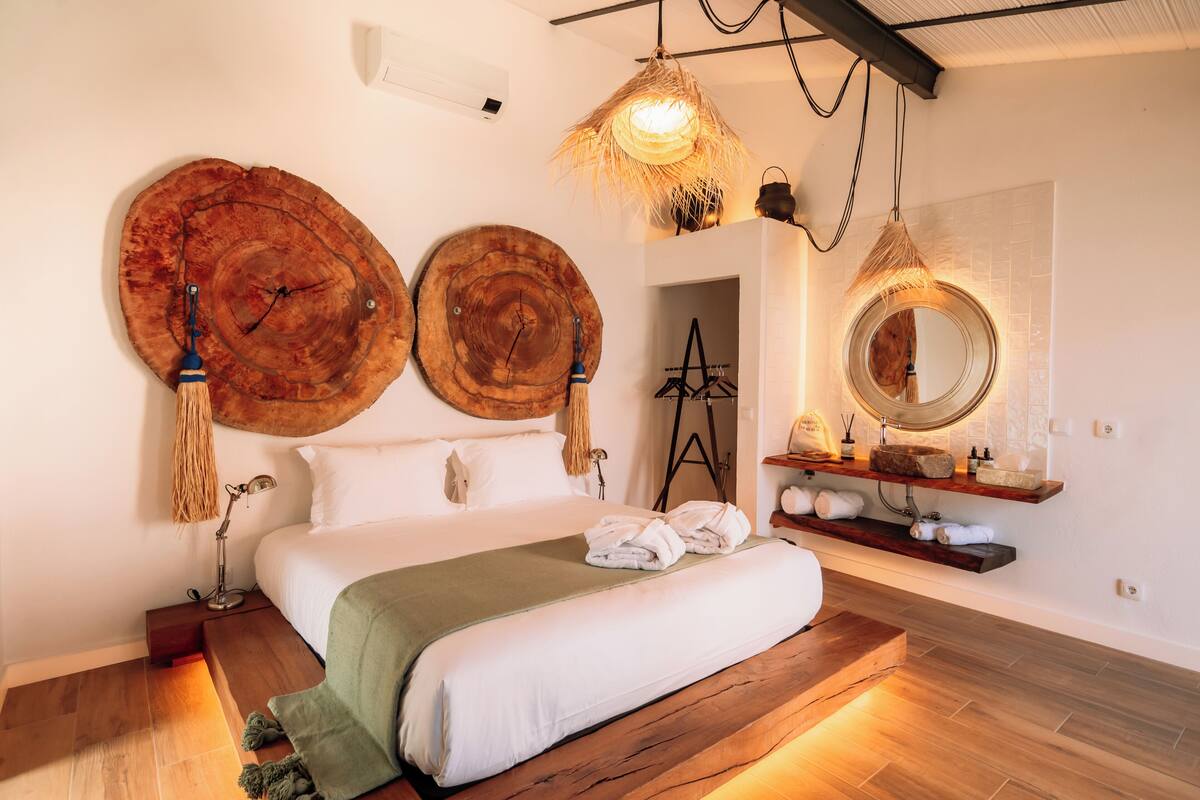 Un dormitorio con piso de madera, cama con ropa de cama blanca, espejo redondo y lámpara colgante.