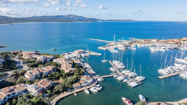 En vy över en kuststad från ovan, med en hamn full av båtar, omgiven av bostadshus och en klar, blå himmel.