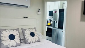 Une chambre avec un lit, des oreillers, un climatiseur fixé au mur, une porte donnant sur un coin cuisine et un téléviseur fixé au mur.