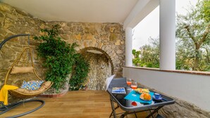 Un pati amb un mur de pedra, una cadira penjant, una taula amb menjar i begudes, i vista als arbres.