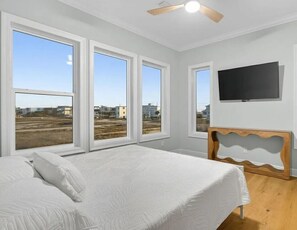 A bedroom with a bed, large windows, a ceiling fan, and a TV mounted on the wall. - Salt Water Heals Everything (Gulf Shores)