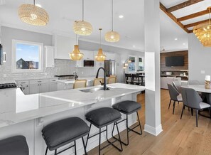 A modern kitchen with white cabinetry, a large island with stools, hanging wicker pendant lights, and a dining area with a TV. - Salt Water Heals Everything (Gulf Shores)