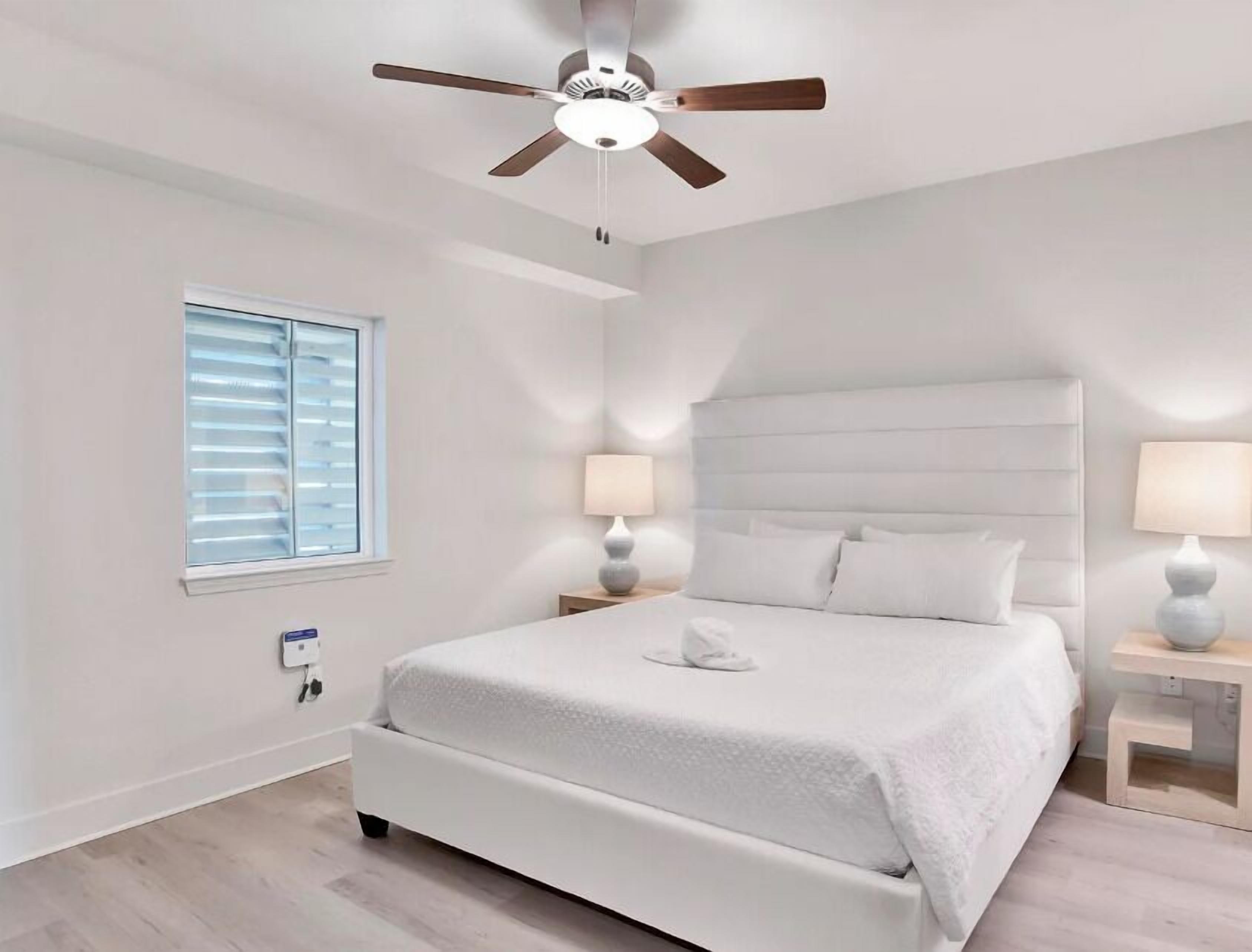 A bedroom with a white bed, a ceiling fan, a window with blinds, and bedside lamps.