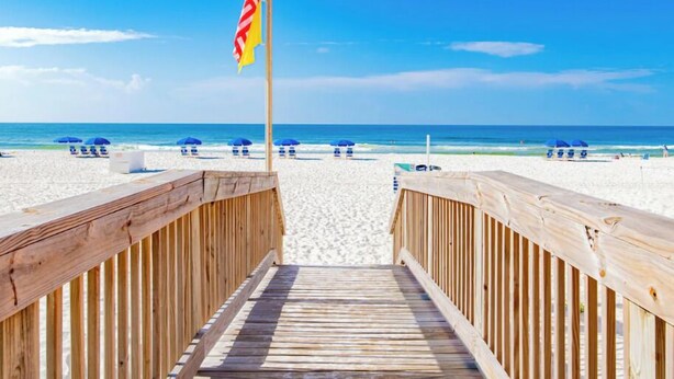 Een houten vlonder die naar een strand leidt, met parasols en een vlag.