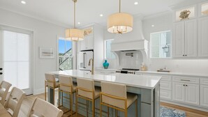 A modern kitchen with white cabinetry, a central island with bar stools, pendant lighting, and a view of the outdoors. - 66 Parks Edge (Orange Beach)