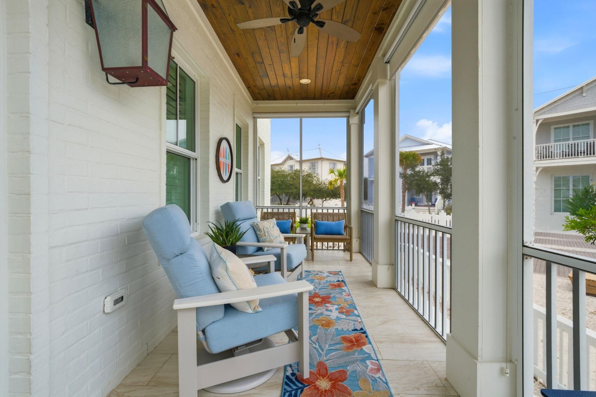 A covered porch with two blue cushioned chairs, a wooden ceiling fan, a white railing, and a view of neighboring houses.
