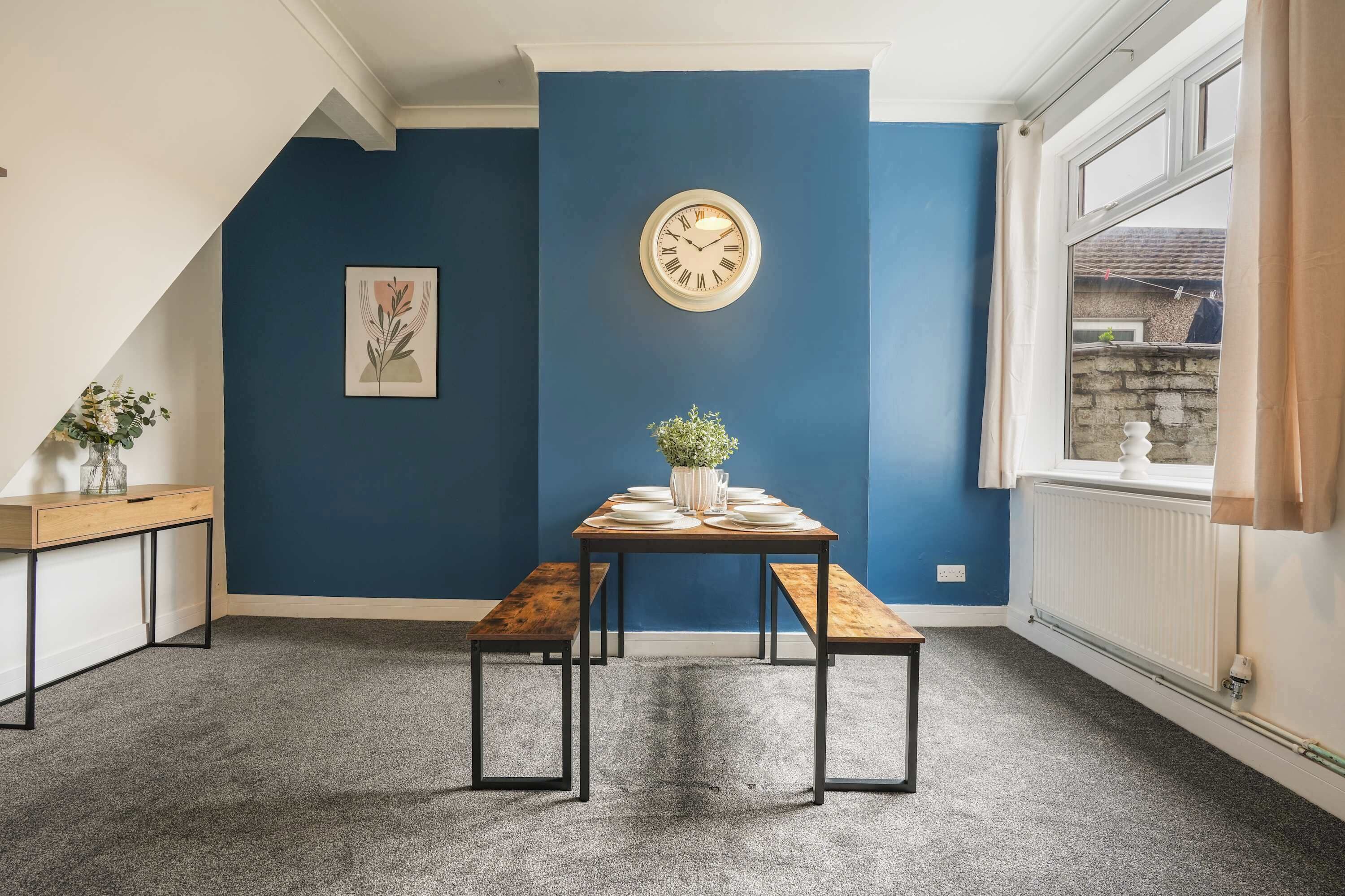 A room with a blue accent wall, a clock, a table with a vase, and benches.