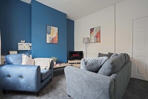 Una sala de estar con una pared azul, un sofá gris, un sillón azul, un televisor mostrando 'NETFLIX' y una lámpara.
