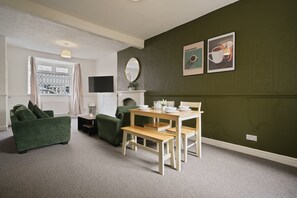A room with a green wall, a white ceiling, a wooden table with chairs, a green sofa, a small white cabinet, and a flat-screen TV mounted on the wall.