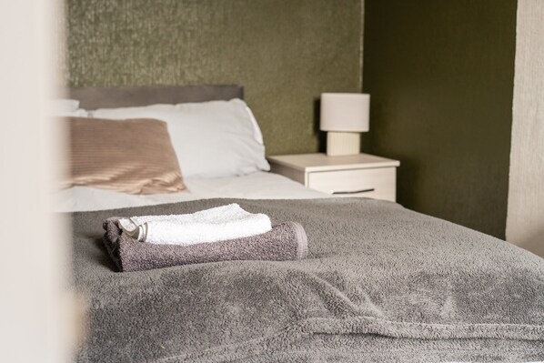 A neatly made bed with a folded towel, a bedside table with a lamp, and a green textured wall.
