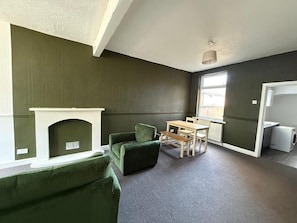 Une pièce avec une cheminée, des fauteuils à revêtement vert, une table en bois avec des chaises, un radiateur et une fenêtre.
