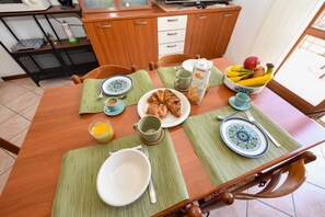 A dining table set with plates, bowls, and a fruit bowl, with a kitchen in the background. - Casa Vuvi - Two-room apartment very close to the beach. (Toscolano Maderno)