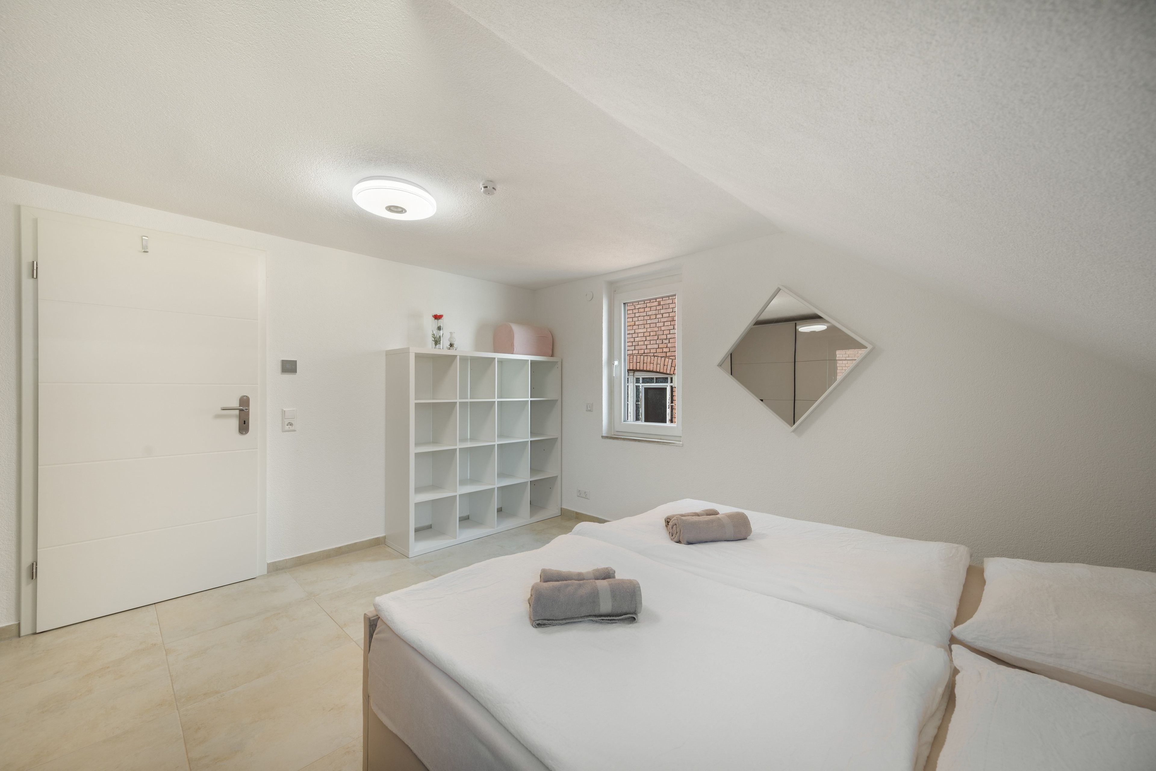 Un dormitorio con cama, armario, espejo y ventana con vista a un muro de ladrillo.