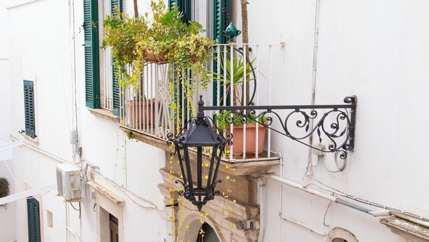 Eine enge Straße mit einem weißen Gebäude, das grüne Fensterläden, einen Balkon mit Blumentöpfen und eine traditionelle Tür zeigt.
