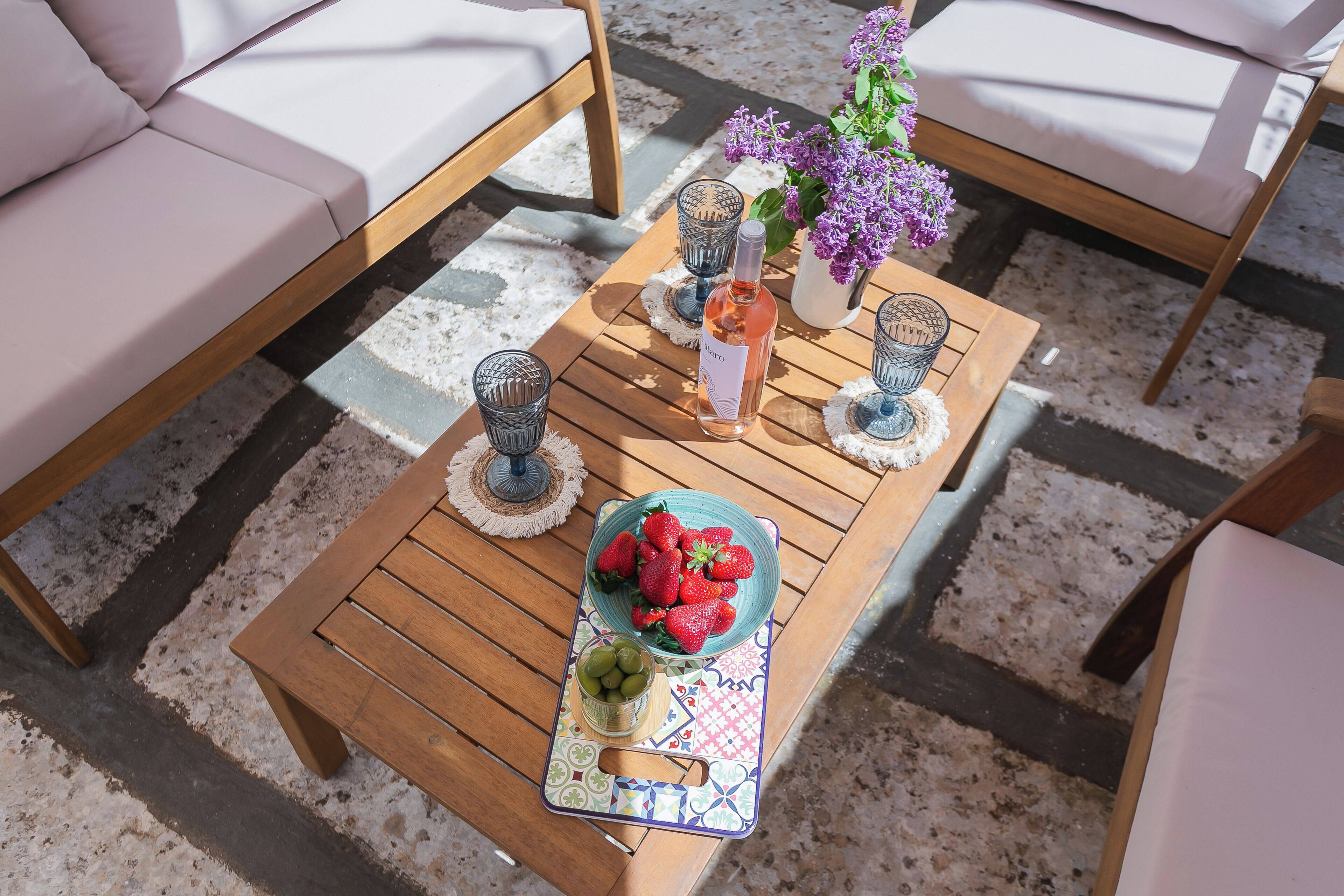 A wooden table with a tray of strawberries and a bottle of wine, surrounded by cushioned chairs and a vase of flowers.