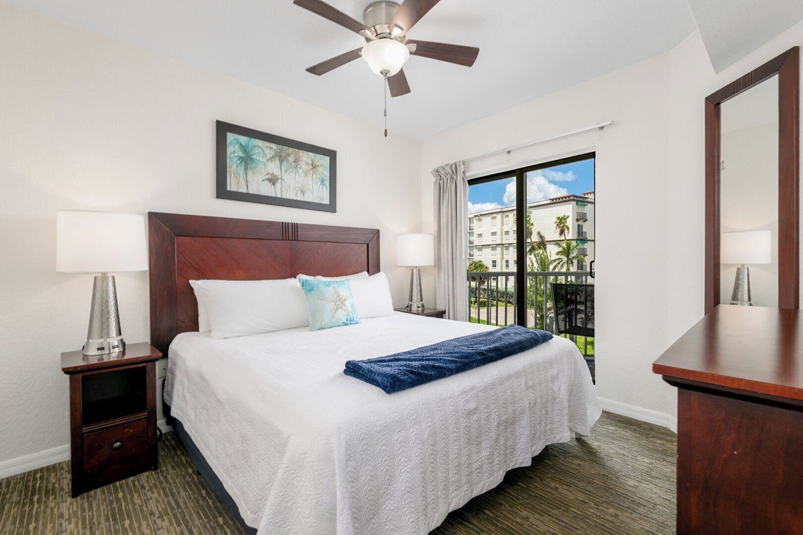 A hotel room with a bed, a ceiling fan, a window with a view, and a nightstand with a lamp.