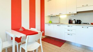 A compact kitchen with white cabinets, a red and white striped wall, a small dining area with white chairs, and a red rug. - Studio Laura - Corte Fasanella (Toscolano Maderno)