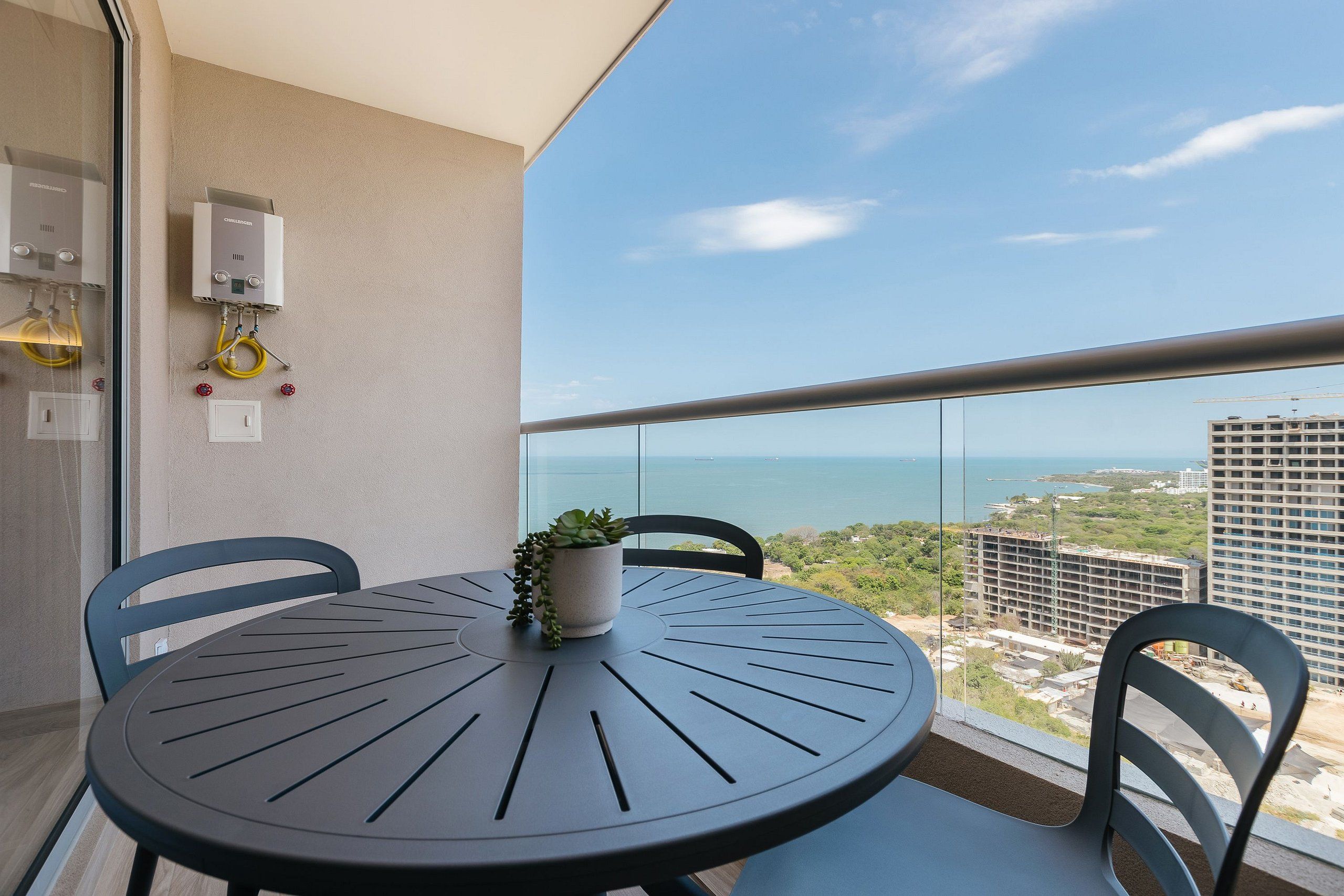 A balcony with a round table, two chairs, a potted plant, and a view of the ocean and buildings.