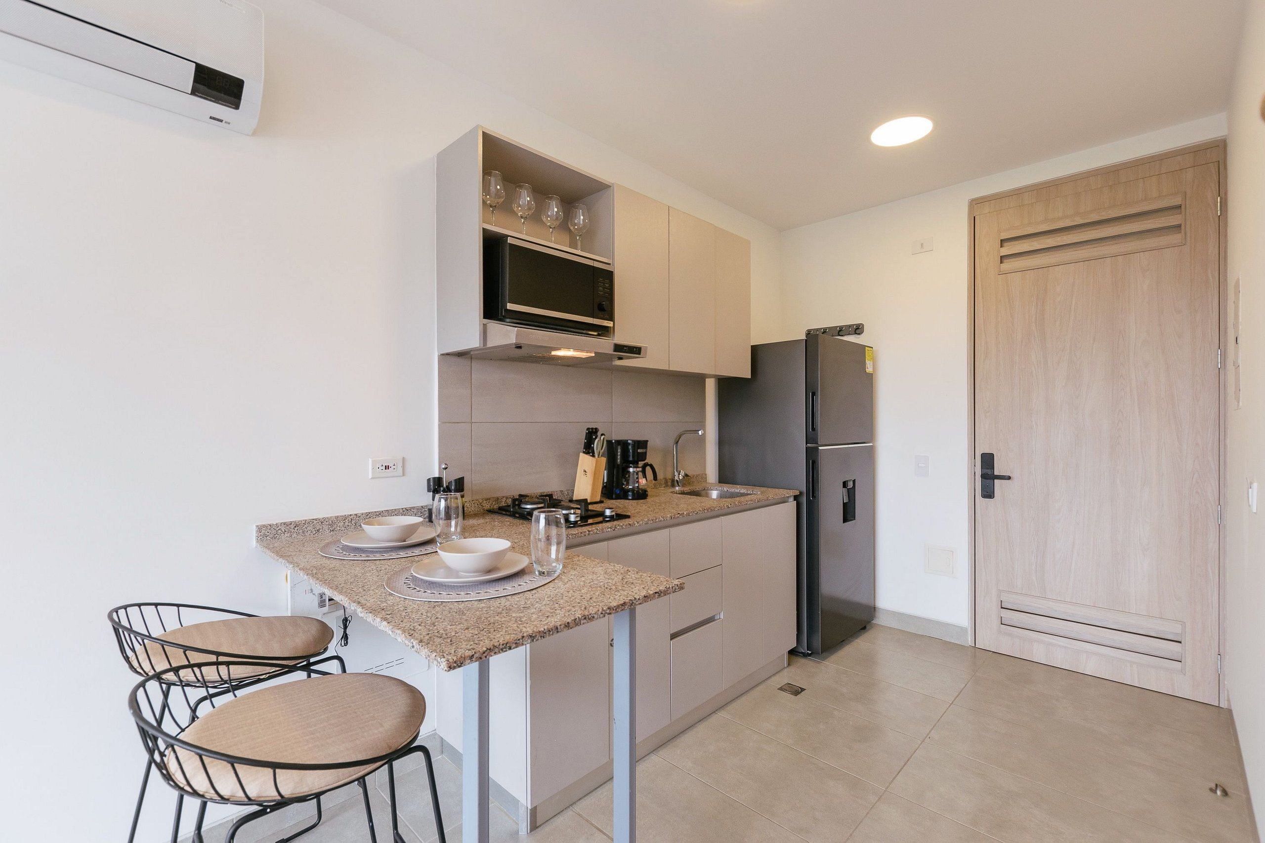 A compact kitchen with a microwave, refrigerator, and a small countertop area. There are two chairs and a shelf with wine glasses.
