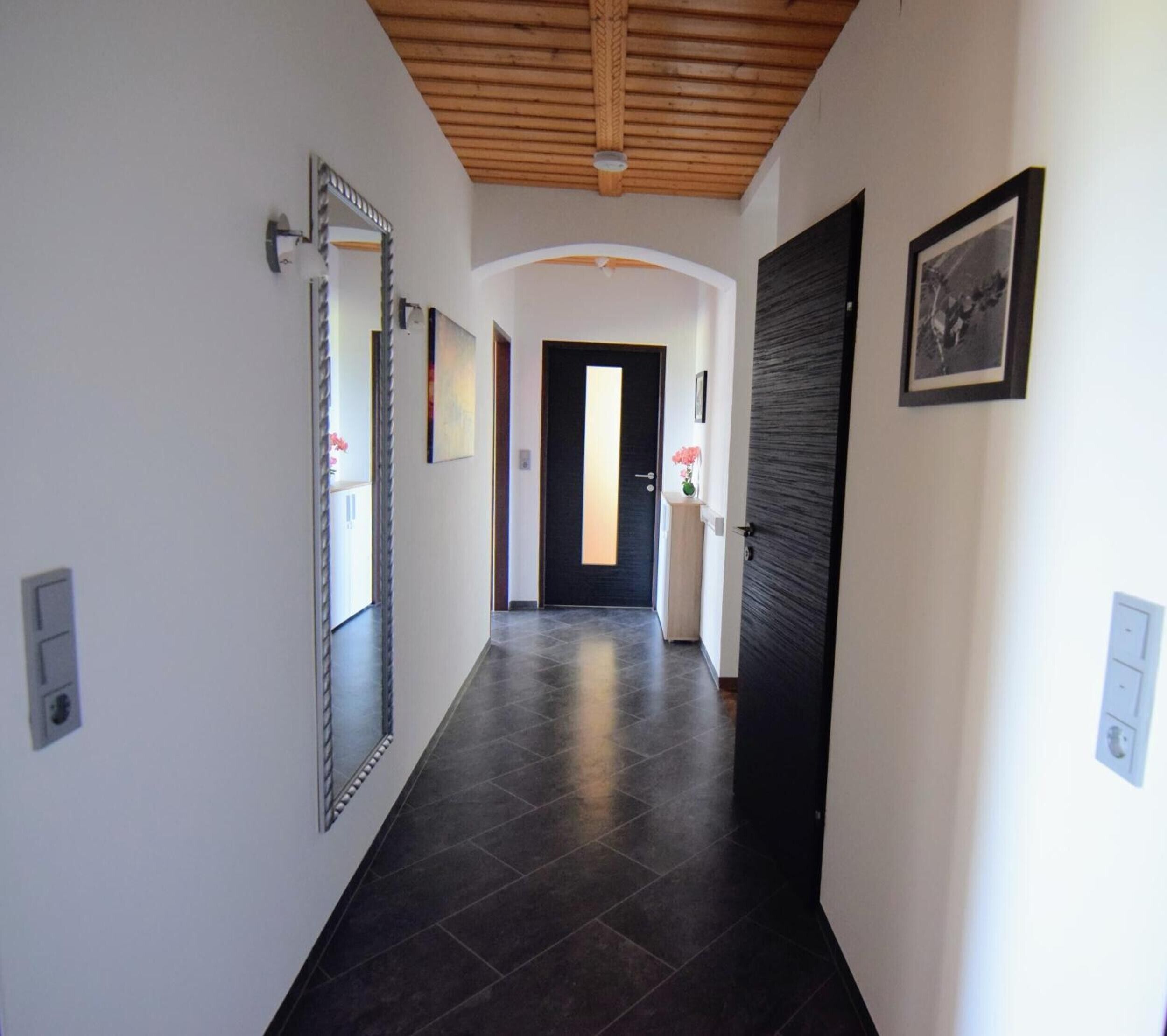 A hallway with a wooden ceiling, dark tiled flooring, a black door, and a mirror on the left wall.