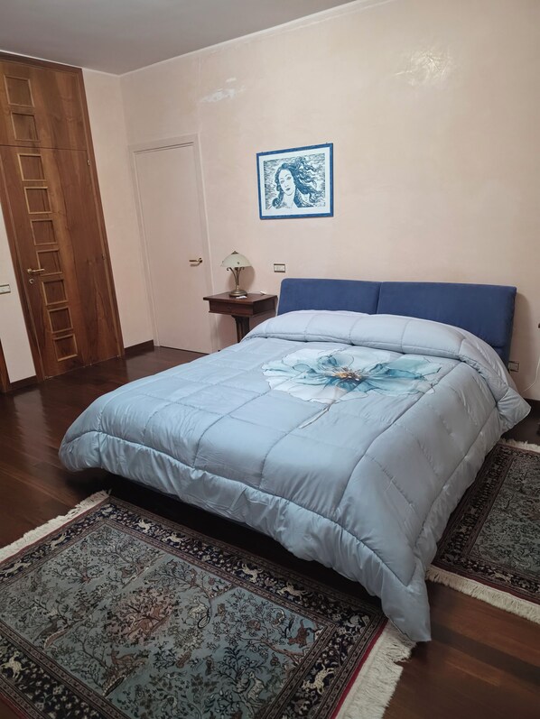 Een slaapkamer met een bed, nachtkastje, lamp, een lijst met afbeelding aan de muur en een deur.