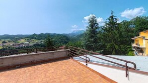 Une terrasse sur le toit, dotée d’une rambarde en métal, d’un sol pavé et offrant une vue sur un paysage verdoyant, avec des arbres et des montagnes à l’horizon.