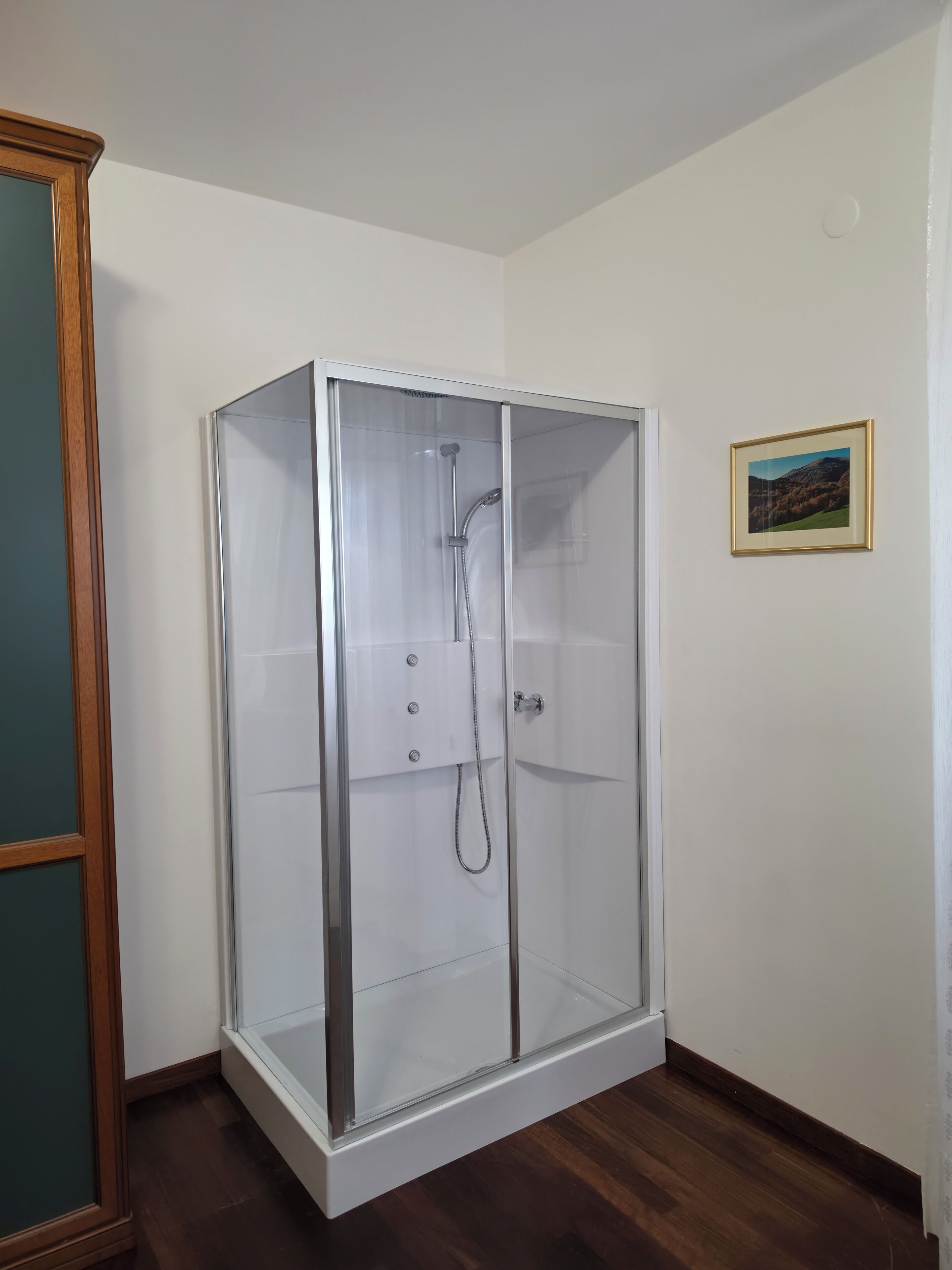 A glass-enclosed shower room with a wooden floor, white walls, and a framed picture on the wall.