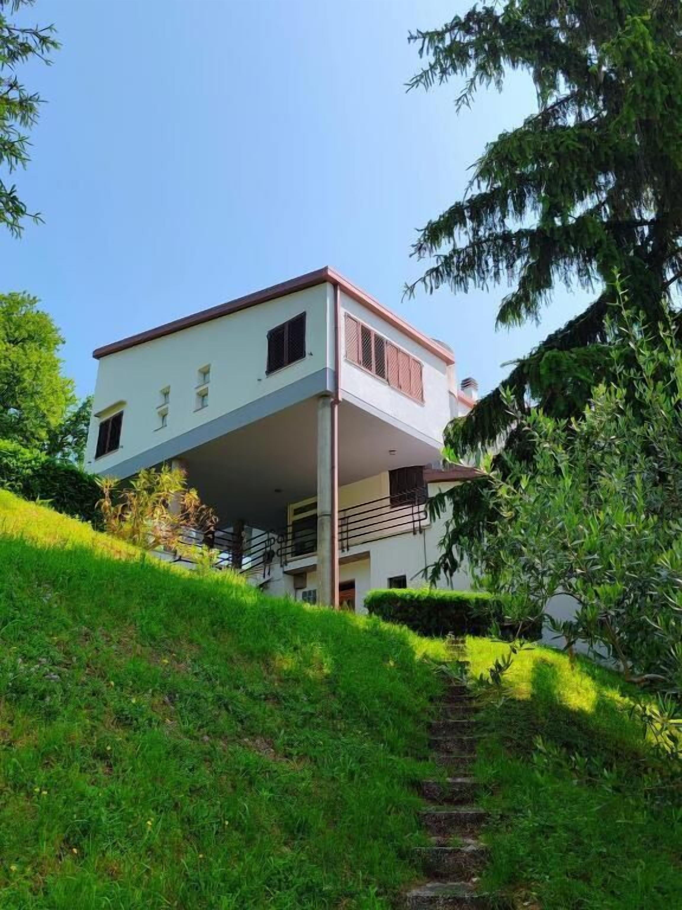 Una casa de dos pisos con balcón, rodeada de vegetación y una pendiente cubierta de césped.