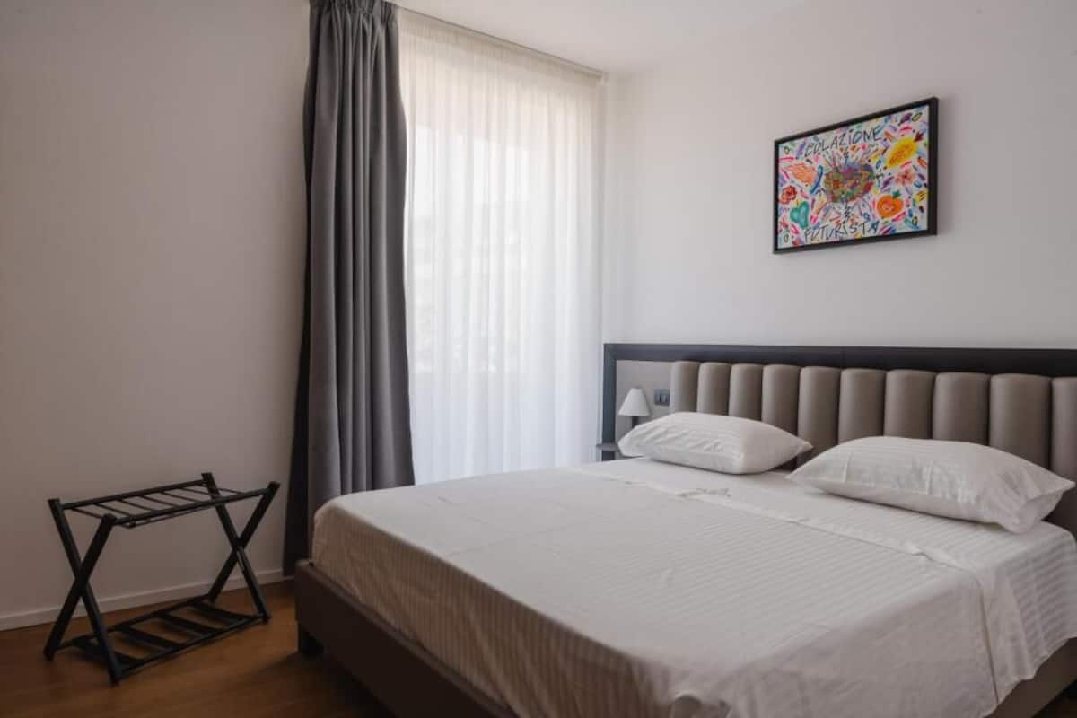 A neatly made bed with white linens, a grey curtain, and a colorful abstract painting on the wall.