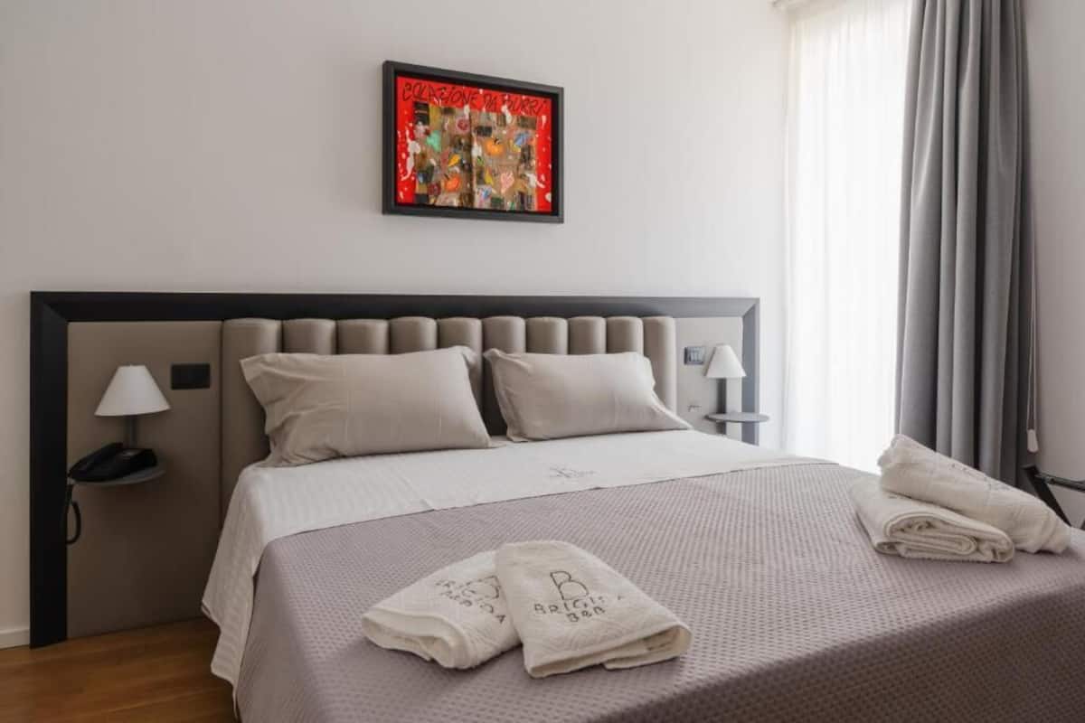 A hotel room with a bed, pillows, a nightstand with a lamp, a framed artwork on the wall, and a window with curtains.