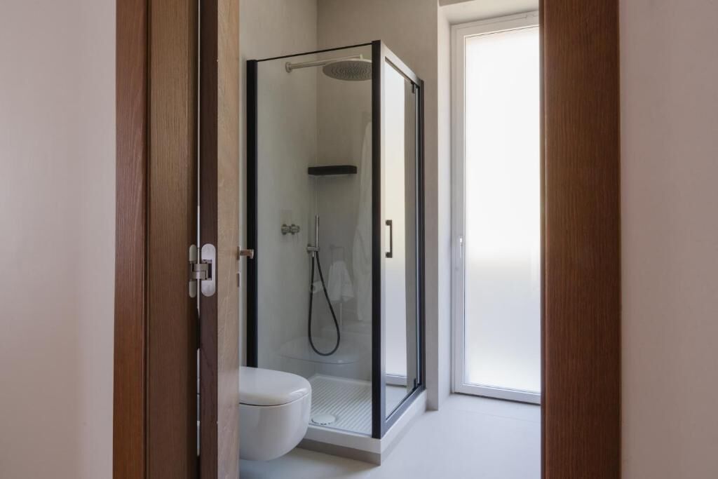 Un bagno moderno con doccia vetrata, un water bianco e una porta che dà su una stanza luminosa.