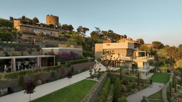 Una villa moderna con jardín, un castillo al fondo y cielo despejado.