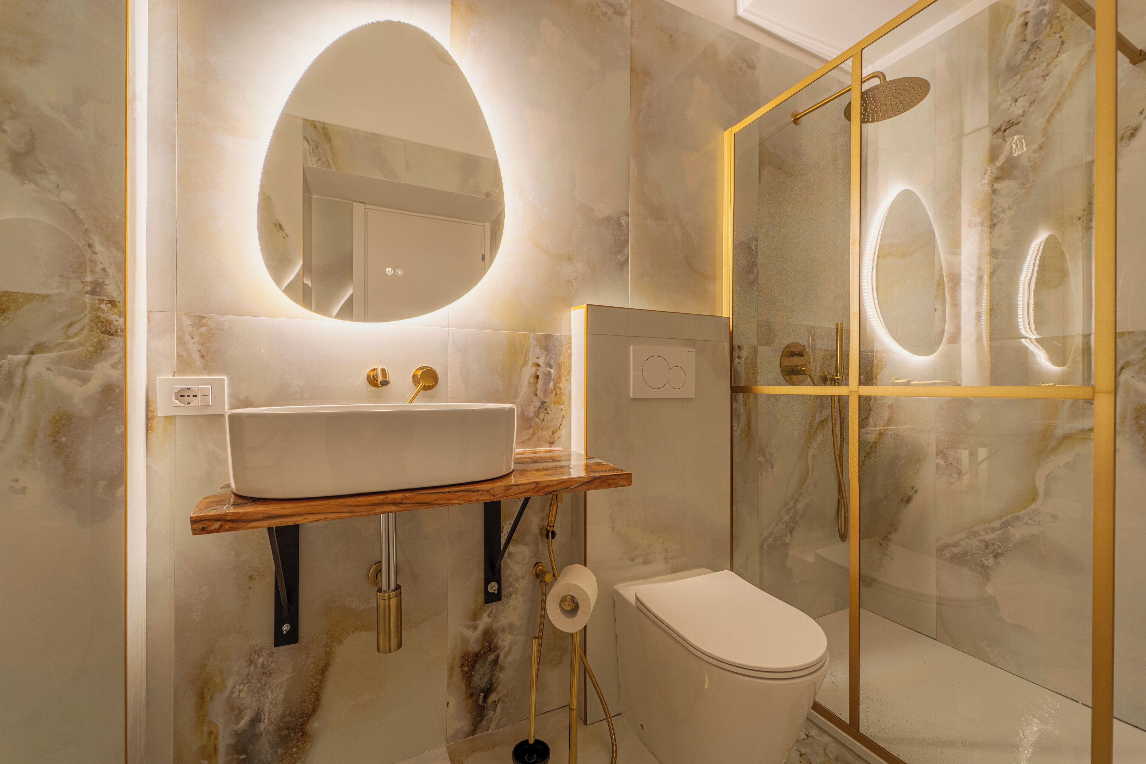 Un bany amb paret de marbre, un tocador de fusta, un lavabo blanc, un vàter i una zona de dutxa amb envàs de vidre.