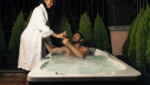 Ein Whirlpool mit zwei Personen, umgeben von Grün und einem Metallzaun.