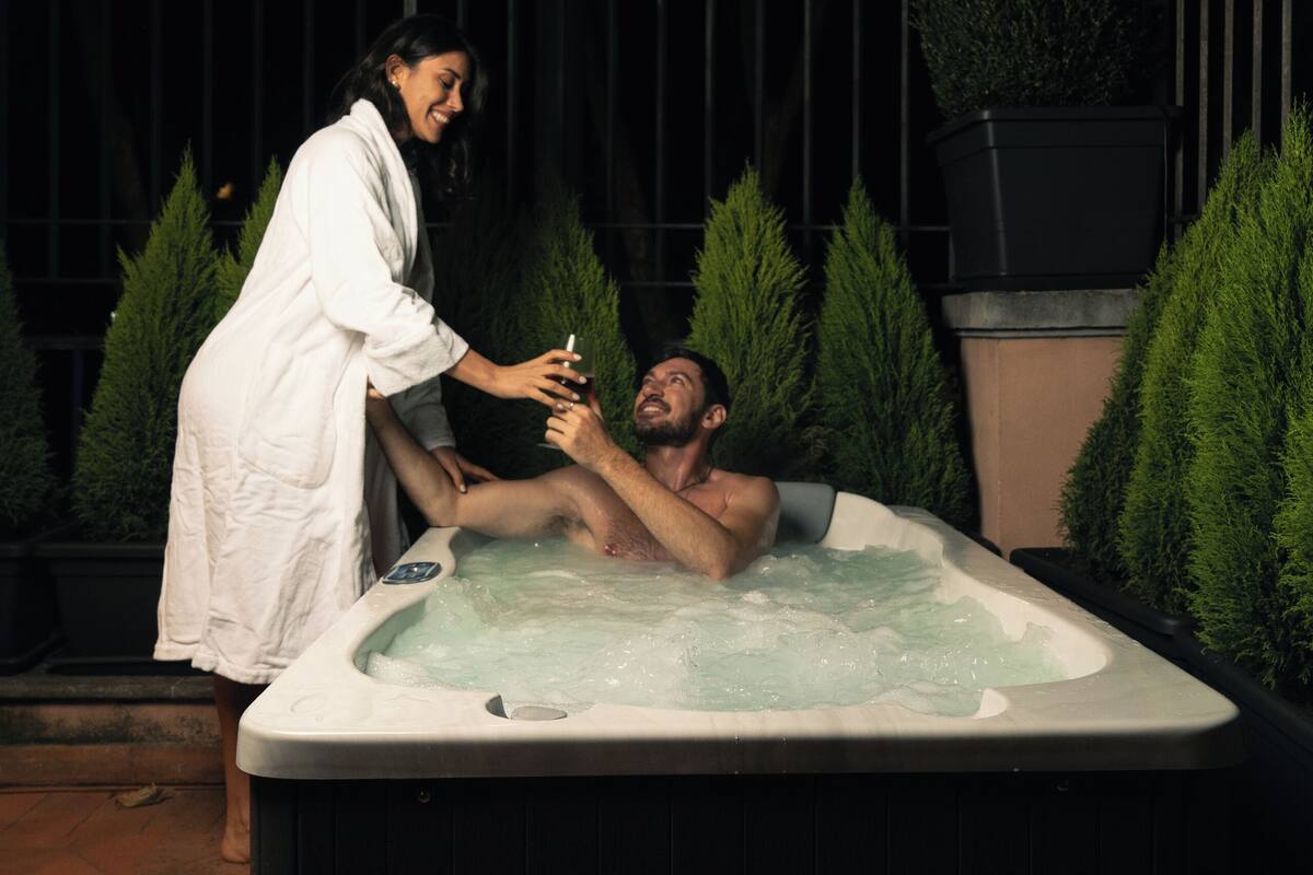 A hot tub with two people, surrounded by greenery and a metal fence.